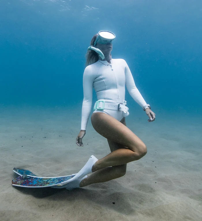 Female freediver kneeling on a sandy ocean floor wearing a white long-sleeve rashguard, white weight belt, dive socks, and colorful patterned fins with a mask on her forehead.