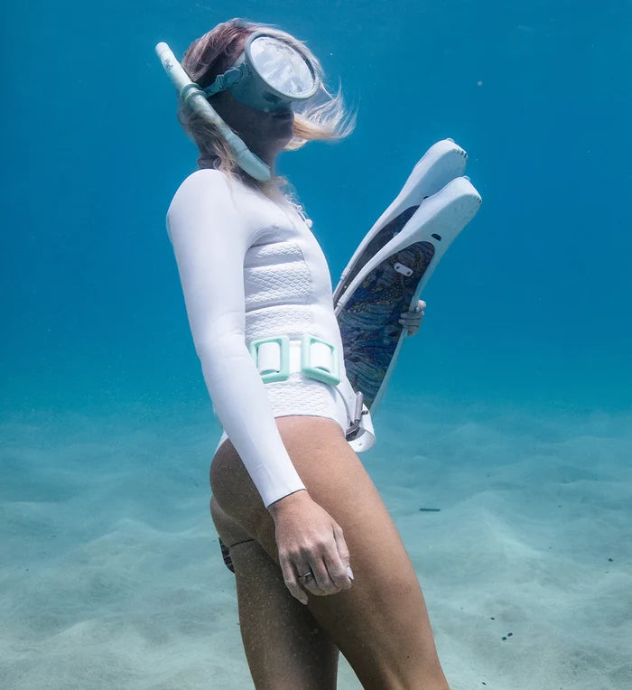 Woman freediver standing on a sandy ocean floor wearing a white long-sleeve rashguard, mint dive weight belt, mask, and snorkel while holding a pair of freediving fins.