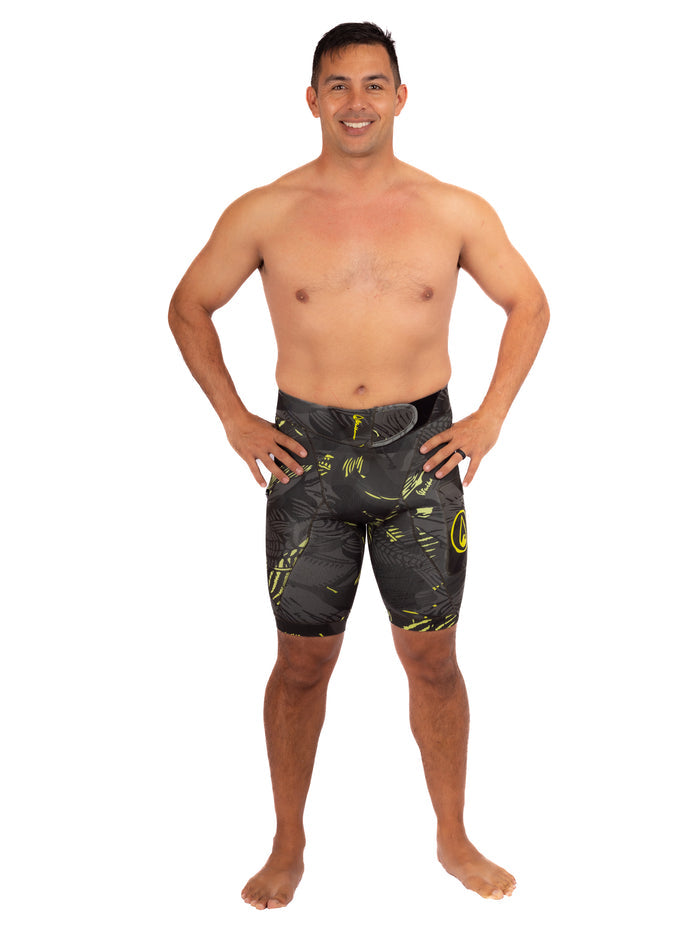Male model wearing Waihana Lime Fishbone Tech Jammers, dark neoprene wetsuit shorts with yellow-green tropical fishbone print and Waihana logo, standing against a white background.