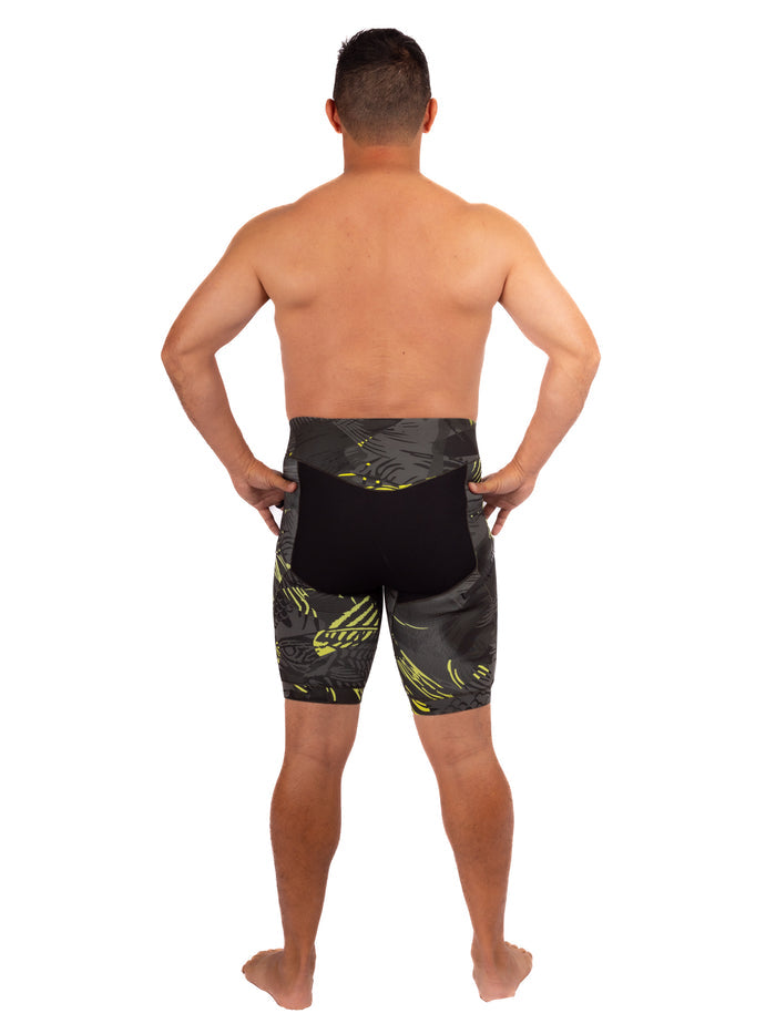 Back view of a male model wearing Waihana Lime Fishbone Tech Jammers featuring a dark grey and yellow-green fishbone tropical print with a black neoprene panel, shot against a white studio background.