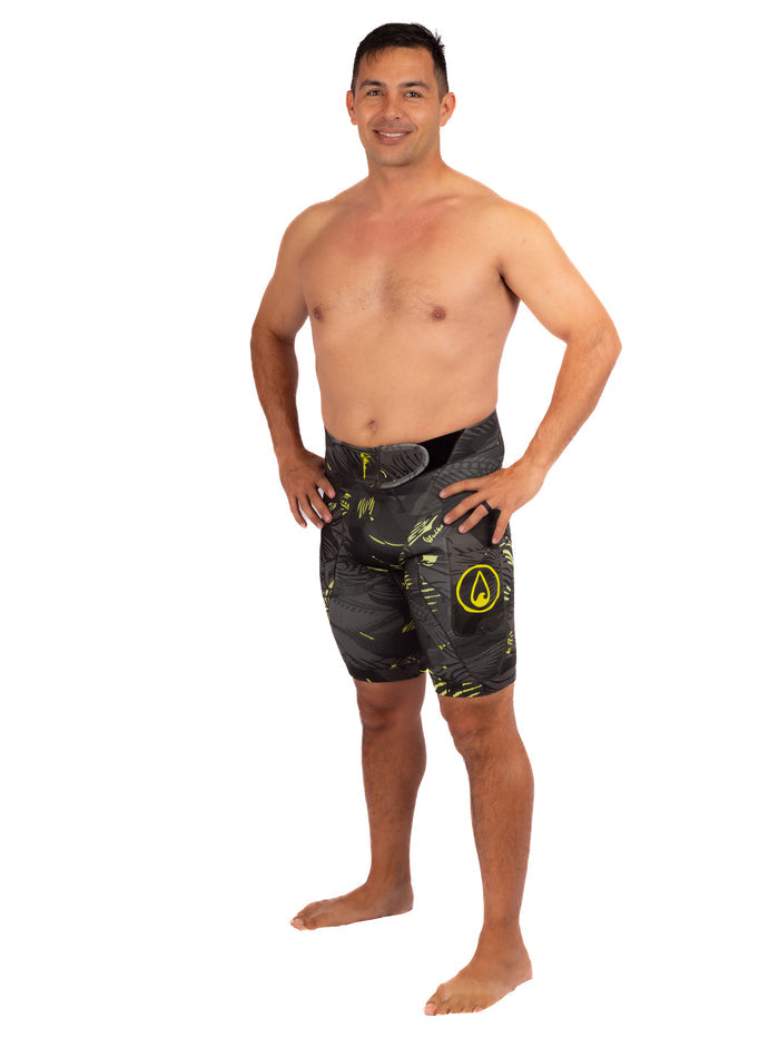 Male model wearing Waihana Lime Fishbone Tech Jammers neoprene wetsuit shorts in a dark fishbone camo pattern with yellow-green accents and Waihana logo, standing shirtless on a white studio background.