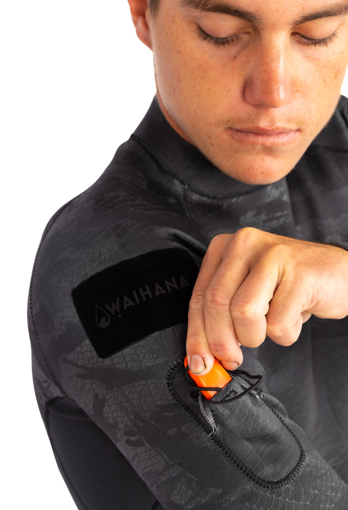 Male model in a Waihana Tropicam Black rear-zip surf rashguard top, demonstrating the wrist zipper with orange pull tab, with the Waihana logo patch visible on the upper arm against a white background.