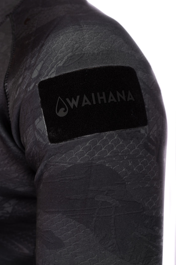 Close-up of the shoulder of the Waihana Men's Tropicam Black Rear-zip Surf Rashguard, showing the embossed fish-scale texture fabric and a black velvet Waihana brand patch on the upper arm.