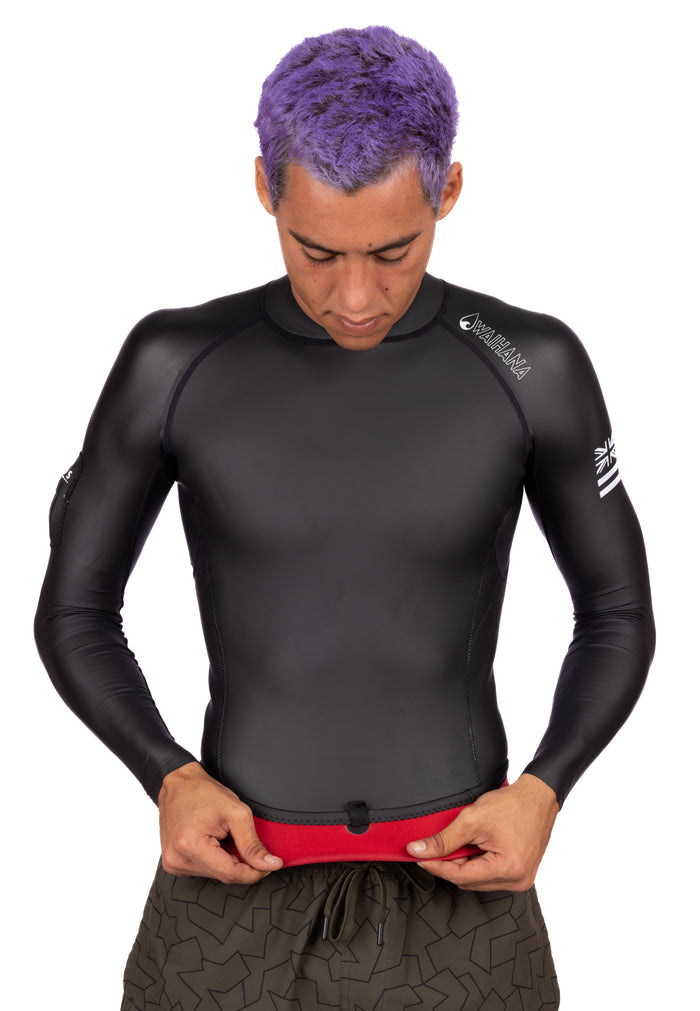 Male model with purple hair wearing a black long-sleeve Waihana WAI-FLEX men's surf impact suit top, adjusting the hem over red board shorts on a white studio background.