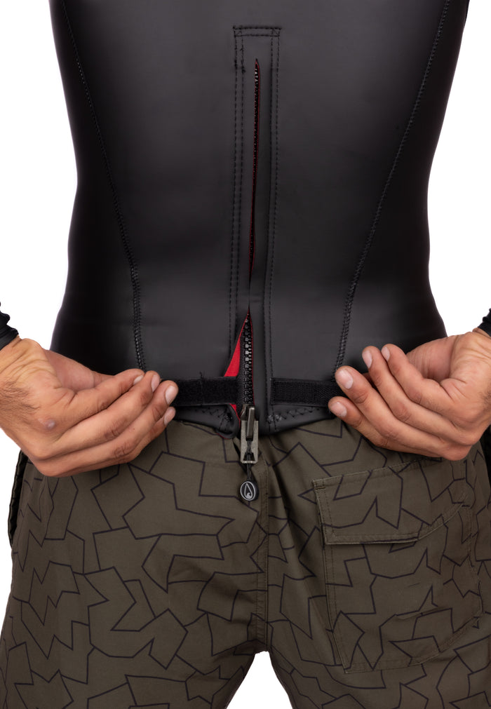 Close-up of a man's hands opening the back zipper of a black WAIHANA WAI-FLEX neoprene surf top with a red interior lining, paired with olive green geometric-print board shorts.