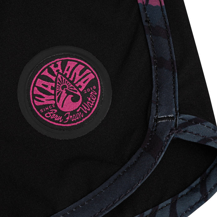Close-up of the Waihana Pink Fishbone W1 Board Shorts featuring an embroidered circular pink Waihana 'Born From Water' logo patch on black fabric, with pink and dark fishbone-patterned lining visible along the seam edge.