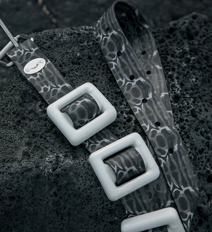 Close-up of the Project Hiu X Bright Buoy Tiger Shark silicone dive weight belt in grey camo pattern with white square buckles, resting on dark volcanic rock.