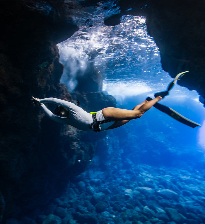 Female freediver in a white rashguard and fins soars gracefully through a sunlit underwater sea cave with vivid blue water and rocky formations below.