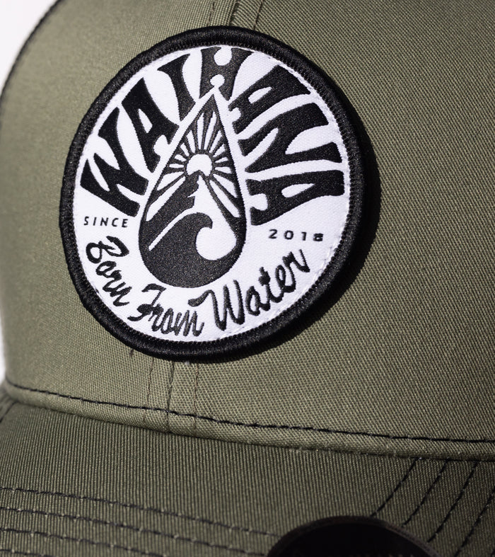 Close-up of the Waihana Ranger Green Mauka Trucker hat showing a circular black and white woven patch reading 'Born From Water Since 2018' on olive green fabric.