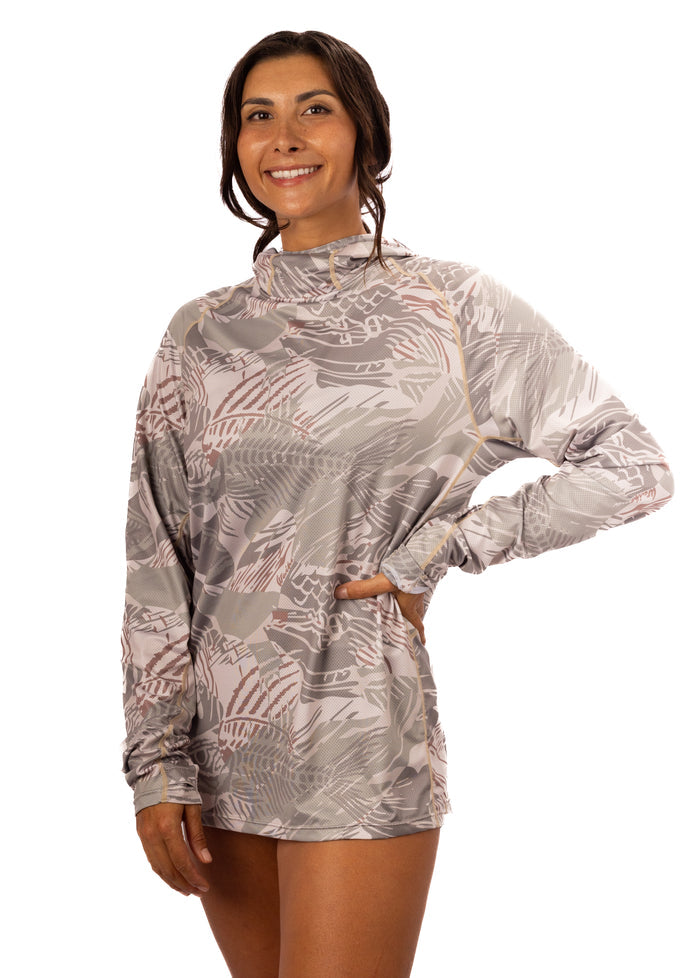Woman smiling and wearing the Waihana Sandy Sun Shirt in Sandy Fishbone print, a long-sleeve hooded sun shirt with a gray and rose tropical leaf pattern, photographed against a white background.