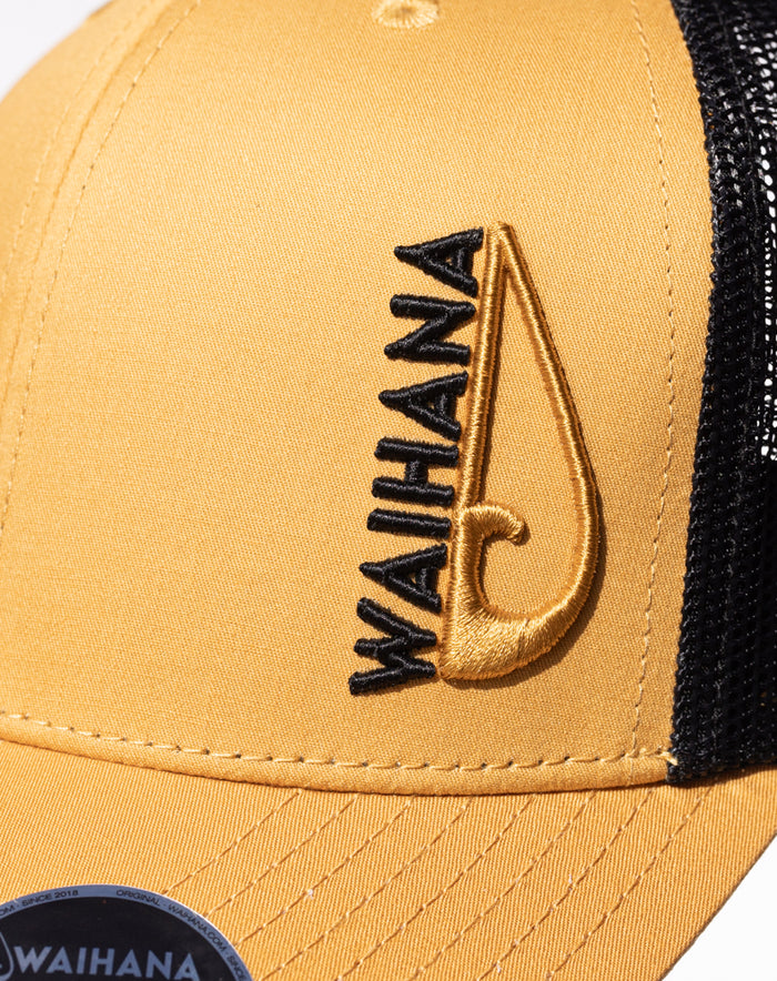 Close-up of a mustard yellow Waihana Sunset Beach Trucker hat with black mesh back panel and embroidered Waihana wordmark alongside a gold fish hook logo.