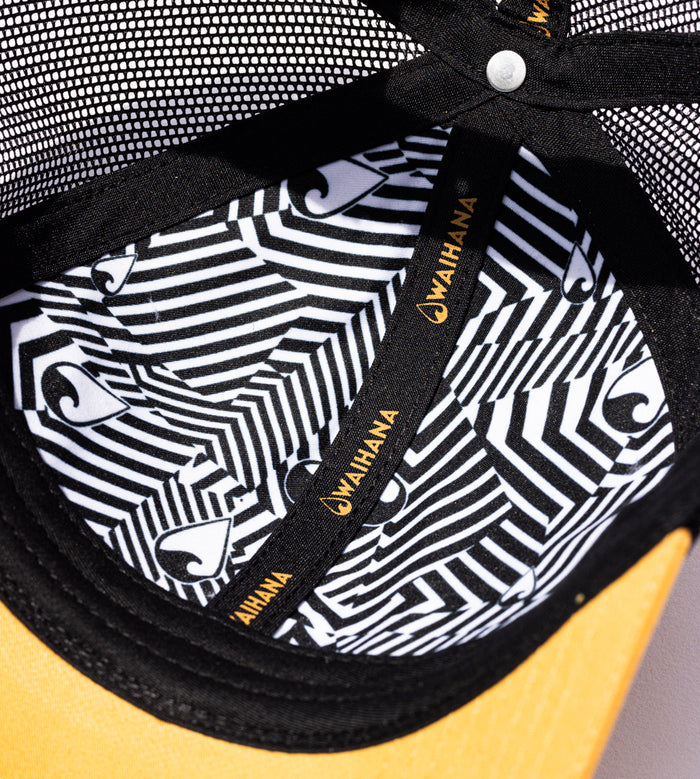 Interior close-up of the Waihana Sunset Beach Trucker hat revealing a striking black-and-white geometric patterned lining with gold Waihana logo labels and a yellow brim.