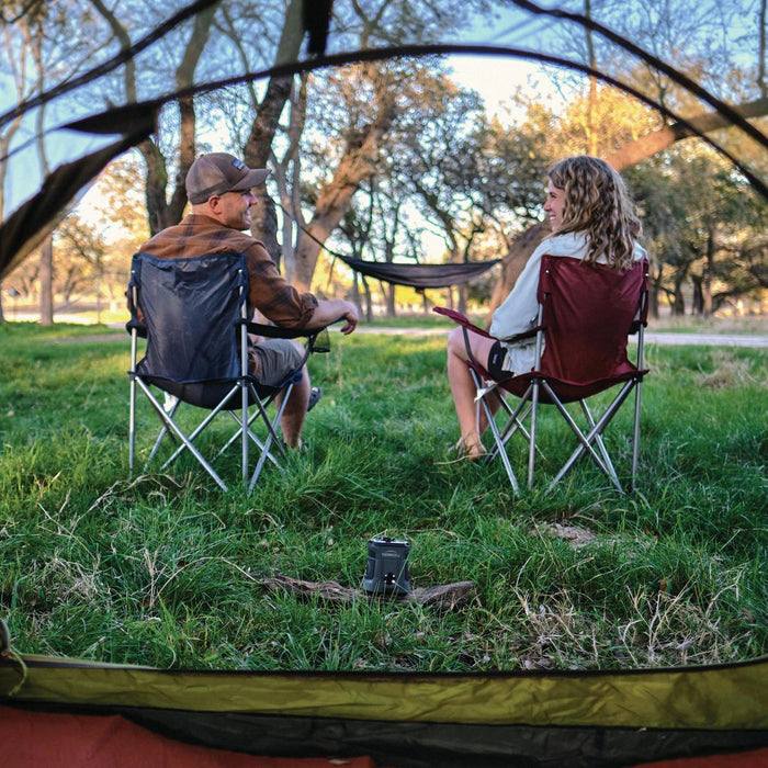 Thermacell EX90 rechargeable mosquito repeller sitting on grass in front of two people camping in folding chairs.