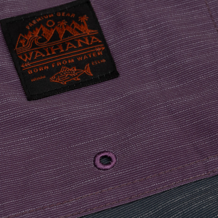 Close-up of Waihana Tidal Moss Cruiser Board Shorts in purple showing the woven 'Born From Water' brand label and embroidered grommet detail on the ripstop fabric.