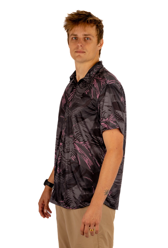 Male model wearing the Waihana Unisex Pink Fishbone Aloha Shirt featuring a dark charcoal and pink tropical fishbone print, paired with khaki shorts, photographed from a side angle on a white background.