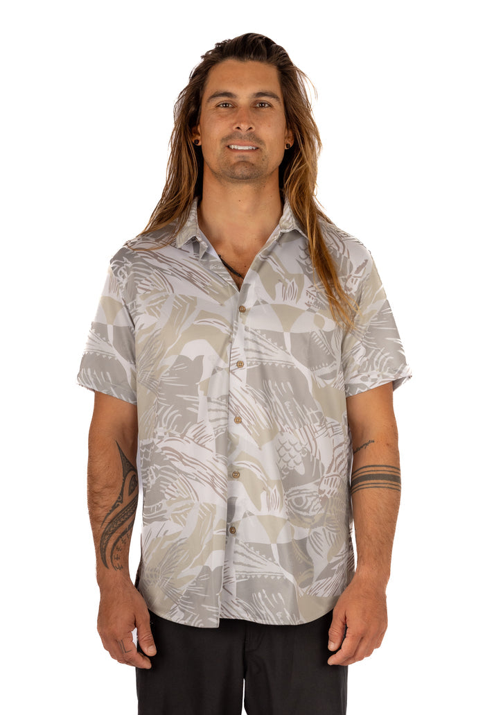 Male model with long hair wearing the Waihana Unisex Sandy Aloha Shirt, a short-sleeve button-up with a grey and tan abstract fish print, shown from the front against a white background.