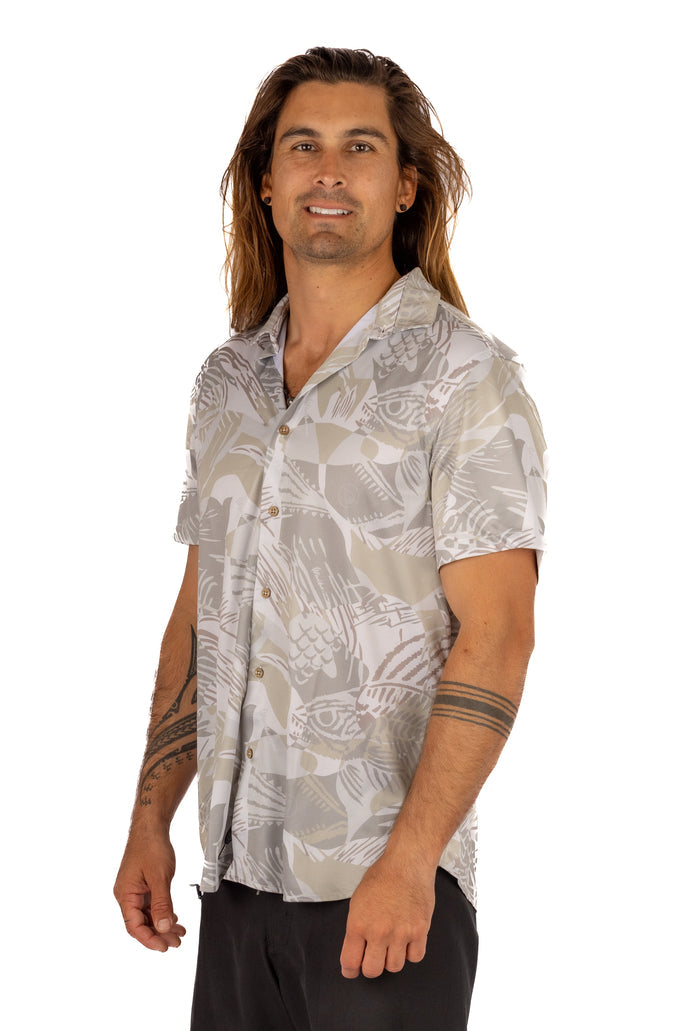Male model with long hair and tattoos wearing the Waihana Unisex Sandy Aloha Shirt featuring an abstract fish print in neutral beige and grey tones, paired with black shorts, on a white background.