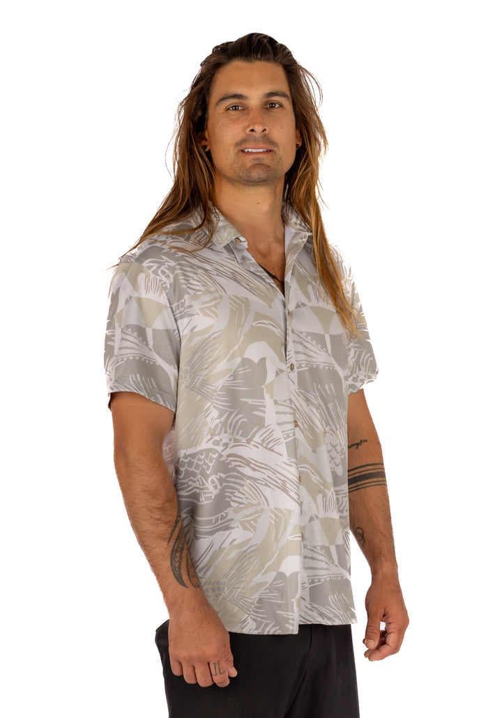 Male model with long hair wearing the Waihana Unisex Sandy Aloha Shirt in a neutral grey and tan abstract tropical print, shown from a three-quarter angle against a white background.