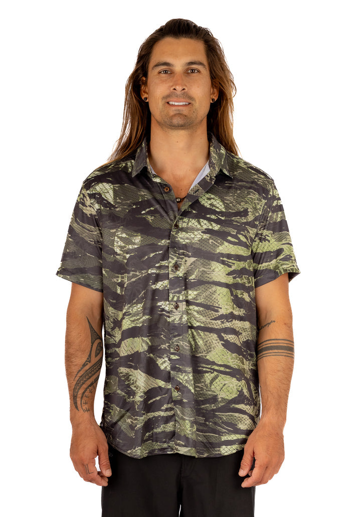 Male model with long hair and tattoos wearing the Waihana Unisex Tropicam Aloha Shirt in a green and charcoal camouflage fish-scale pattern, shown from the front against a white background.