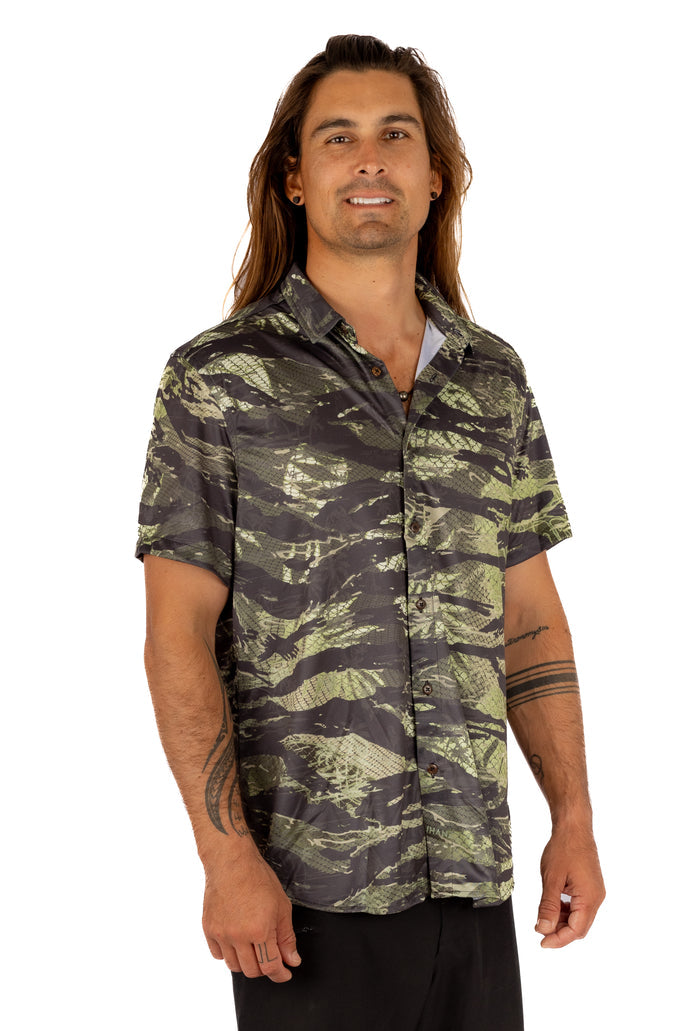 Male model with long hair wearing the Waihana Unisex Tropicam Aloha short-sleeve button-up shirt in green camouflage print, shown from the front against a white background.