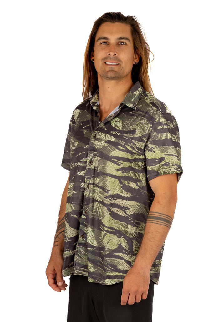 Male model with long hair wearing a Waihana Unisex Tropicam Aloha Shirt in green and grey camouflage print with short sleeves and button front, shown from a three-quarter angle on a white background.