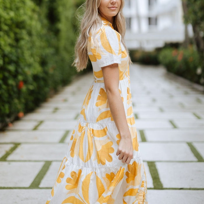 Woman wearing a white and yellow floral print ankle-length button-up dress with a collar in a resort setting.
