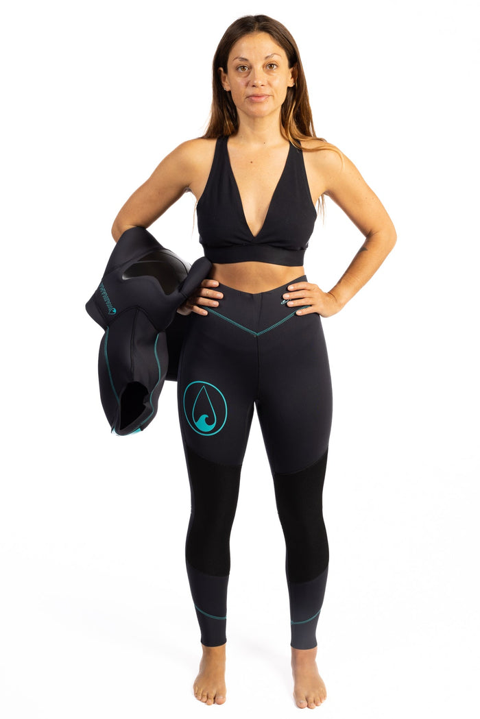 Woman modeling Waihana Women's Essentials black neoprene wetsuit pants with teal stitching and Waihana logo, holding the matching wetsuit jacket under her arm against a white studio background.