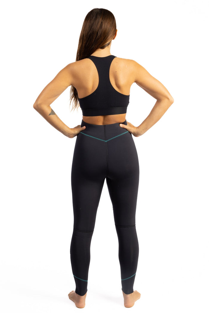 Back view of a woman wearing Waihana Women's Essentials black neoprene wetsuit pants with teal accent stitching and a black racerback top, on a white studio background.