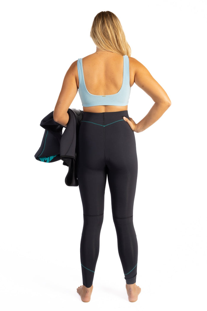Back view of a woman wearing Waihana Women's Essentials black neoprene wetsuit pants with teal accent stitching and a light blue sports bra, holding a wetsuit top, on a white studio background.
