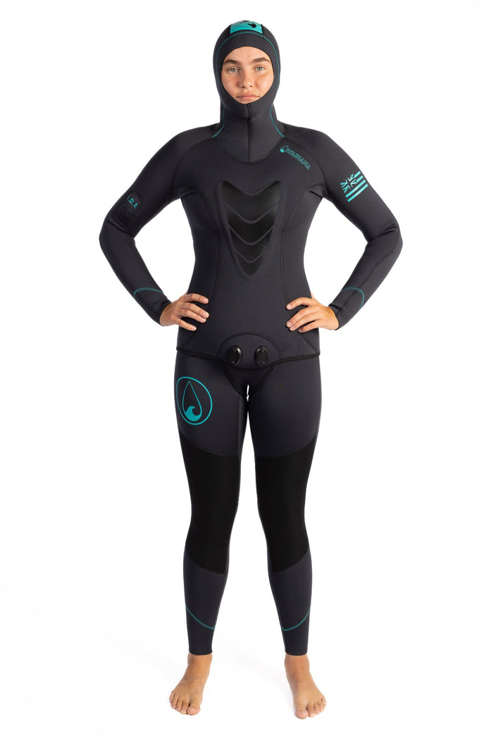 Female model wearing a black Waihana Women's Essentials two-piece wetsuit with integrated hood and teal accent stitching, standing with hands on hips against a white background.