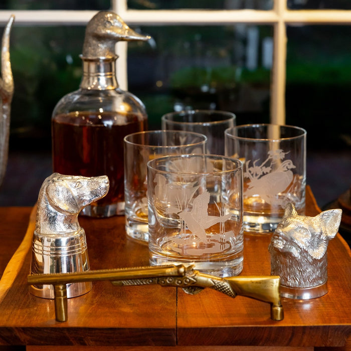 Wooden charcuterie tray with brass shotgun handles, featuring etched wildlife glassware and silver animal-head barware.